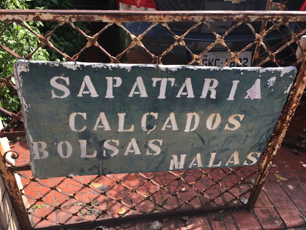 A weathered metal sign is affixed to a rusted fence, with the words 'Sapataria Calçados Bolsas Malas' in faded white paint. The background is a dark green color, with visible chipping and peeling. Behind the fence is a parked car with a visible license plate and green foliage on the left side.