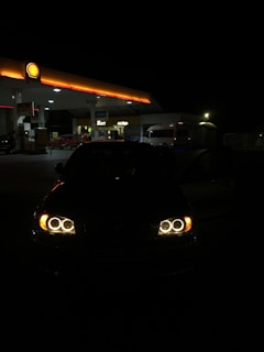 A dimly lit gas station is visible at night with its illuminated signage and canopy. In the foreground, a car with its headlights on is partially visible. The gas station, branded with a Shell logo, is surrounded by darkness, highlighting the warm glow of the lights.