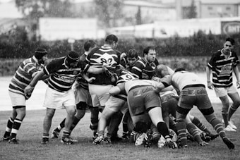 A group of rugby players are engaged in a scrum on a wet field, with raindrops visible. They are wearing striped jerseys and shorts, and some players have headgear for protection. The scene captures the intensity and physicality of the sport.