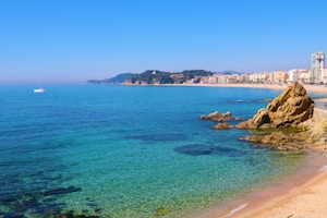 A serene coastal landscape featuring clear turquoise waters meeting the sandy shore. Rugged rocks are visible in the foreground, adding texture to the scene. A collection of modern buildings lines the coastline in the background. A white sailboat is gently floating in the distance, with green hills framing the horizon.