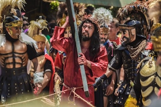 A group of people are dressed in elaborate costumes, resembling soldiers and a figure in a red robe with a crown of thorns holding a wooden beam. The costumes feature feathers, metallic accents, and dark armor-like details, suggesting a historical or theatrical reenactment.