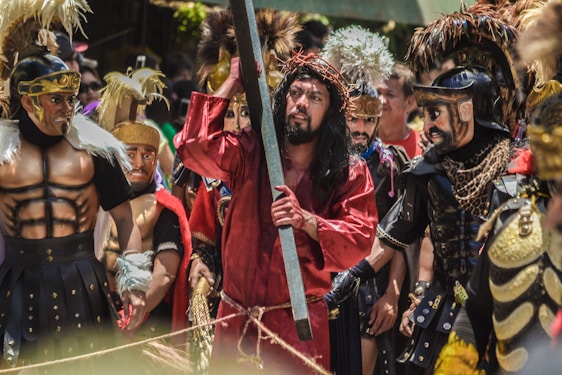 A group of people are dressed in elaborate costumes, resembling soldiers and a figure in a red robe with a crown of thorns holding a wooden beam. The costumes feature feathers, metallic accents, and dark armor-like details, suggesting a historical or theatrical reenactment.