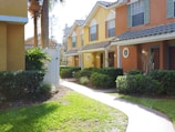 A row of colorful townhouses with yellow and orange facades, framed by manicured bushes and grass along a winding pathway. The houses feature tiled roofs, large windows with shutters, and small round windows near the entry doors. Bright natural light suggests a sunny day, and palm trees are visible in the background.