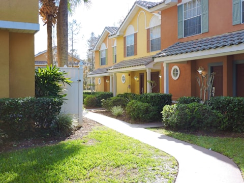 A row of colorful townhouses with yellow and orange facades, framed by manicured bushes and grass along a winding pathway. The houses feature tiled roofs, large windows with shutters, and small round windows near the entry doors. Bright natural light suggests a sunny day, and palm trees are visible in the background.