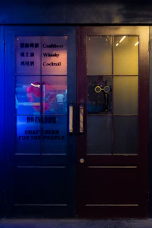 A pair of dark wooden doors with glass panels and text advertising craft beer, whisky, and cocktails. The left door features some text in both English and another language, with the phrases 'Craftbeer', 'Whisky', 'Cocktail', and 'BREWDOG' written on it. A warm yellow light shines through the glass of the right door, hinting at indoor lighting. The left side is illuminated with a blue light, adding a vibrant contrast to the scene.