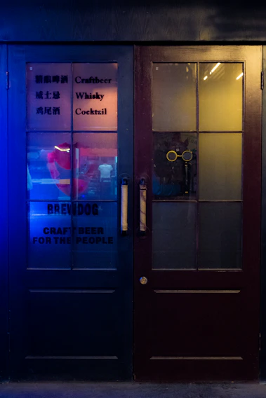 A pair of dark wooden doors with glass panels and text advertising craft beer, whisky, and cocktails. The left door features some text in both English and another language, with the phrases 'Craftbeer', 'Whisky', 'Cocktail', and 'BREWDOG' written on it. A warm yellow light shines through the glass of the right door, hinting at indoor lighting. The left side is illuminated with a blue light, adding a vibrant contrast to the scene.