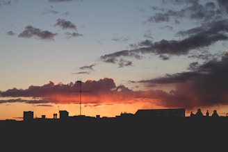 Silhouette of a city skyline at sunset with a prominent cross structure surrounded by orange and dark clouds. The horizon displays darkened building outlines against a gradient sky transitioning from orange to blue.