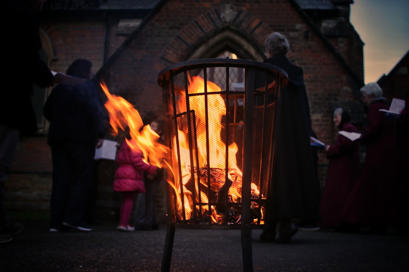 A metal fire basket in the foreground contains bright, orange flames flickering upwards. In the background, several people are gathered around, dressed in warm clothing and holding papers, possibly singing or participating in an outdoor event near a brick building with arched windows.