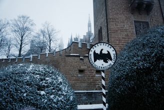 A snow-covered scene featuring a section of a brick castle wall with battlements. The foreground shows dense bushes dusted with snow, and a circular sign with the words 'Preussen Hohenzollernsche Lande' and a black eagle at the center. In the background, leafless trees and the spire of a building can be seen through a gloomy, overcast sky.