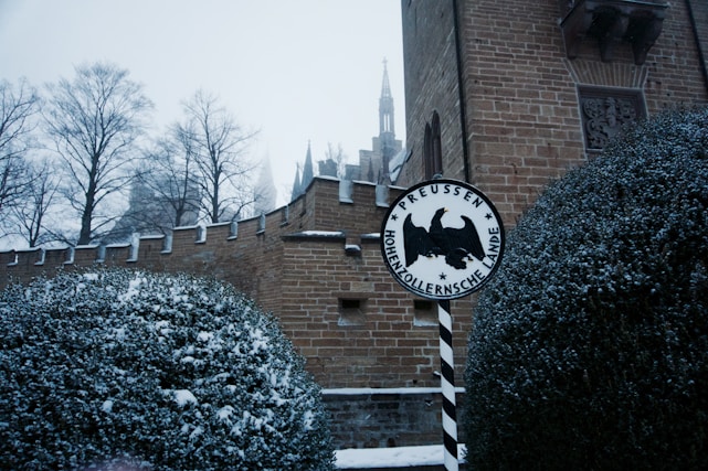 A snow-covered scene featuring a section of a brick castle wall with battlements. The foreground shows dense bushes dusted with snow, and a circular sign with the words 'Preussen Hohenzollernsche Lande' and a black eagle at the center. In the background, leafless trees and the spire of a building can be seen through a gloomy, overcast sky.