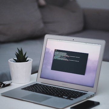 A sleek MacBook Air laptop displaying code on its screen is placed on a white table. Next to it, there's a small potted succulent plant in a white pot. In the background, a grey couch is visible. On the table, there's also a smartphone and a pair of sunglasses.