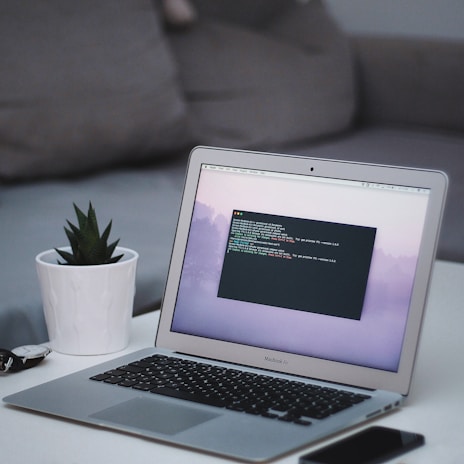 A sleek MacBook Air laptop displaying code on its screen is placed on a white table. Next to it, there's a small potted succulent plant in a white pot. In the background, a grey couch is visible. On the table, there's also a smartphone and a pair of sunglasses.