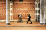A musician stands under a decorated brick archway, playing a guitar. The blurred figure of a pedestrian is captured walking past the musician, adding a sense of motion to the scene. The background features an ornate brick and stone wall with inscriptions.