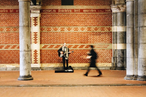 A musician stands under a decorated brick archway, playing a guitar. The blurred figure of a pedestrian is captured walking past the musician, adding a sense of motion to the scene. The background features an ornate brick and stone wall with inscriptions.