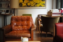 A cozy and inviting interior space with a brown leather armchair in the foreground. The room features wooden flooring and is adorned with a variety of armchairs and bookshelves. A large yellow and green abstract painting hangs on the wall, adding a touch of artistic flair. Books are stacked on a wooden table, contributing to the atmosphere of a relaxed reading nook.
