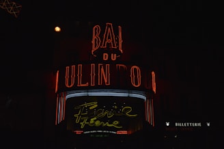 A vibrant neon sign displays the words 'Moulin Rouge' in red and a cursive 'Féerie' in yellow, set against a dark background. The sign is illuminated with additional red and white lights, evoking a lively and classic nightlife atmosphere. The words 'BILLETTERIE' and 'TICKET OFFICE' are visible, suggesting a place to purchase tickets.
