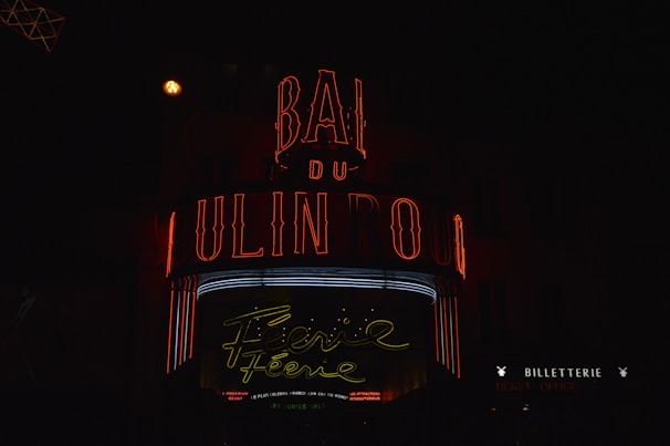 A vibrant neon sign displays the words 'Moulin Rouge' in red and a cursive 'Féerie' in yellow, set against a dark background. The sign is illuminated with additional red and white lights, evoking a lively and classic nightlife atmosphere. The words 'BILLETTERIE' and 'TICKET OFFICE' are visible, suggesting a place to purchase tickets.