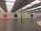 A large, industrial gallery space with a high ceiling featuring metal beams and skylights. There are four large, solid-colored square paintings on the white wall: red, black, yellow, and blue, from left to right. The floor is polished concrete, and there is a circle of light-colored stones arranged on the floor in the foreground to the right.