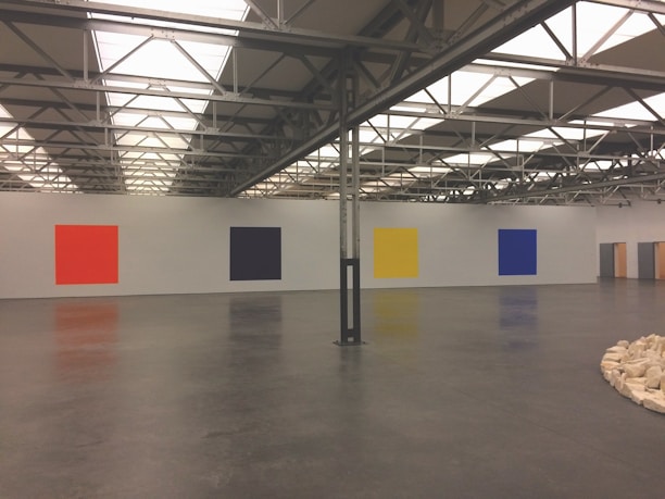 A large, industrial gallery space with a high ceiling featuring metal beams and skylights. There are four large, solid-colored square paintings on the white wall: red, black, yellow, and blue, from left to right. The floor is polished concrete, and there is a circle of light-colored stones arranged on the floor in the foreground to the right.