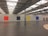 A large, industrial gallery space with a high ceiling featuring metal beams and skylights. There are four large, solid-colored square paintings on the white wall: red, black, yellow, and blue, from left to right. The floor is polished concrete, and there is a circle of light-colored stones arranged on the floor in the foreground to the right.