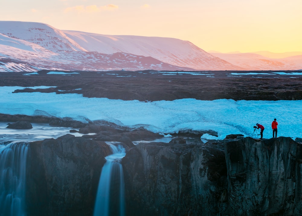 two persons standing on cliff with waterfall during daytime