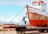 A weathered fishing boat rests on a metal platform with two people working on it. The boat's hull shows signs of rust and age. In the background, there are snow-capped mountains under a partly cloudy sky, exhibiting a scenic landscape. One person stands on a ladder painting or repairing the front of the boat, while another sits nearby, appearing to be taking a break.