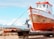 A weathered fishing boat rests on a metal platform with two people working on it. The boat's hull shows signs of rust and age. In the background, there are snow-capped mountains under a partly cloudy sky, exhibiting a scenic landscape. One person stands on a ladder painting or repairing the front of the boat, while another sits nearby, appearing to be taking a break.