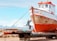 A weathered fishing boat rests on a metal platform with two people working on it. The boat's hull shows signs of rust and age. In the background, there are snow-capped mountains under a partly cloudy sky, exhibiting a scenic landscape. One person stands on a ladder painting or repairing the front of the boat, while another sits nearby, appearing to be taking a break.