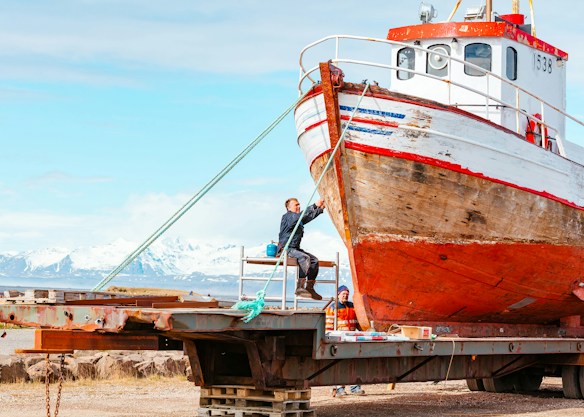 A weathered fishing boat rests on a metal platform with two people working on it. The boat's hull shows signs of rust and age. In the background, there are snow-capped mountains under a partly cloudy sky, exhibiting a scenic landscape. One person stands on a ladder painting or repairing the front of the boat, while another sits nearby, appearing to be taking a break.