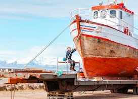 A weathered fishing boat rests on a metal platform with two people working on it. The boat's hull shows signs of rust and age. In the background, there are snow-capped mountains under a partly cloudy sky, exhibiting a scenic landscape. One person stands on a ladder painting or repairing the front of the boat, while another sits nearby, appearing to be taking a break.