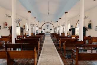 A well-lit church interior with wooden pews aligned on both sides of a central aisle leading to an altar. The walls are decorated with religious statues and framed pictures. Large white columns support the high ceiling, and several ceiling fans are visible. Stained glass windows and wall-mounted lights contribute to the serene atmosphere.