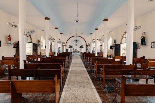 A well-lit church interior with wooden pews aligned on both sides of a central aisle leading to an altar. The walls are decorated with religious statues and framed pictures. Large white columns support the high ceiling, and several ceiling fans are visible. Stained glass windows and wall-mounted lights contribute to the serene atmosphere.