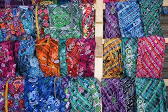 An array of brightly colored, intricately patterned fabrics displayed in rows. Each fabric features unique designs, including floral patterns and geometric shapes, using a vibrant palette. The arrangement showcases a diverse mix of colors and textures.