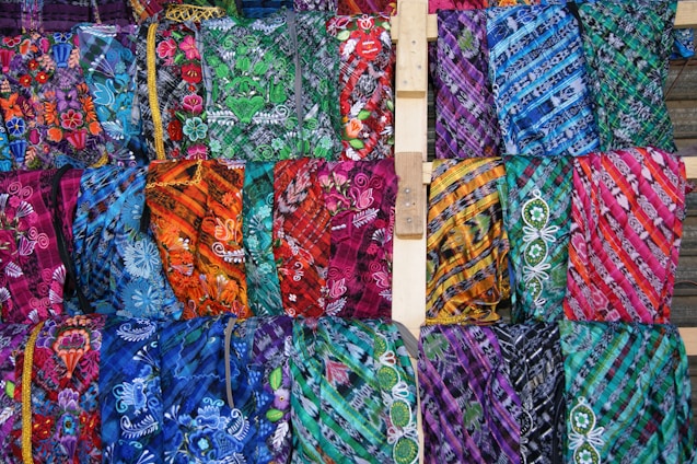 An array of brightly colored, intricately patterned fabrics displayed in rows. Each fabric features unique designs, including floral patterns and geometric shapes, using a vibrant palette. The arrangement showcases a diverse mix of colors and textures.