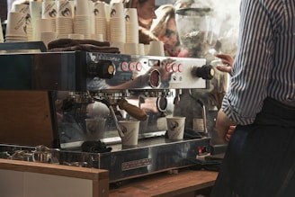 A barista is operating a large, commercial espresso machine making coffee. Numerous coffee cups are stacked on top, and steam is rising from the machine. The setting seems busy with visible reflections of people and activity in the background.