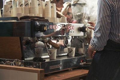 A barista is operating a large, commercial espresso machine making coffee. Numerous coffee cups are stacked on top, and steam is rising from the machine. The setting seems busy with visible reflections of people and activity in the background.