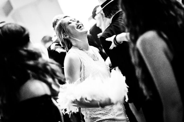 A black and white image capturing a joyful, lively moment in what appears to be a social gathering or party. A woman is in the center, laughing and wearing a sparkling dress, surrounded by others who are interacting closely. The scene conveys motion and energy.