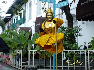 A street performer is dressed in a vibrant yellow costume with a metallic mask, sitting cross-legged on a pole above ground, giving an impression of levitation. The performer is surrounded by a railing and lush green plants in a lively outdoor setting. In the background, there is a white building with green shutters and signage pointing to museums.