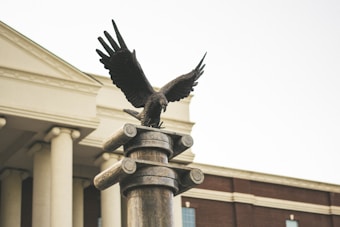 A bronze eagle statue with its wings spread wide is perched on top of a cylindrical column. The background features a classic building with white pillars and a red brick facade.