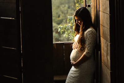 A pregnant woman in a lace dress gently cradles her belly as she stands by a wooden doorway with sunlight streaming in. The background features lush greenery, creating a serene and warm atmosphere.