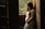 A pregnant woman in a lace dress gently cradles her belly as she stands by a wooden doorway with sunlight streaming in. The background features lush greenery, creating a serene and warm atmosphere.