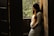 A pregnant woman in a lace dress gently cradles her belly as she stands by a wooden doorway with sunlight streaming in. The background features lush greenery, creating a serene and warm atmosphere.