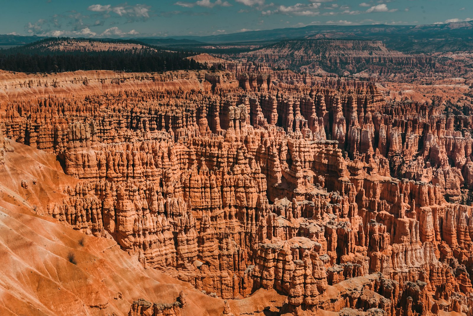 A high-angle landscape photograph showing a vast amphitheater of orange-red hoodoos and rock formations under a blue sky with clouds.