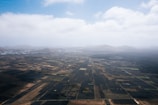 An aerial view of a vast agricultural landscape featuring a patchwork of dark and light fields stretching towards distant mountains under a partly cloudy sky.