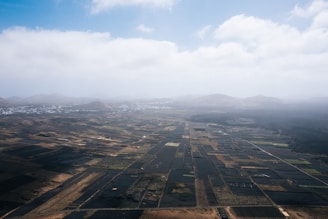 An aerial view of a vast agricultural landscape featuring a patchwork of dark and light fields stretching towards distant mountains under a partly cloudy sky.