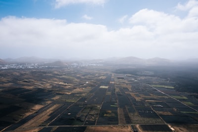 An aerial view of a vast agricultural landscape featuring a patchwork of dark and light fields stretching towards distant mountains under a partly cloudy sky.