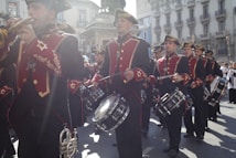 A marching band in formal attire performs in a street parade. The band members wear red and black uniforms with gold accents and are playing brass instruments and drums. Sunlight casts a dramatic flare across the scene, enhancing the atmosphere.