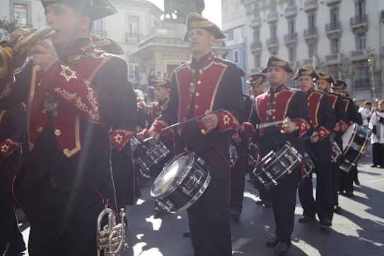 A marching band in formal attire performs in a street parade. The band members wear red and black uniforms with gold accents and are playing brass instruments and drums. Sunlight casts a dramatic flare across the scene, enhancing the atmosphere.