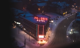 A nighttime winter scene featuring a neon-lit hotel situated on a snowy street corner. The hotel's sign glows with red and blue lights, contrasting against the dark and cold surroundings. Snow covers the rooftops and streets, with sparse streetlights illuminating the winding road.