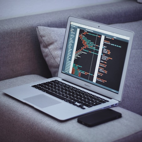 A laptop sits on a gray fabric couch, displaying code on its screen. Next to it, a smartphone lies flat. The code appears to be written in HTML or a similar programming language.